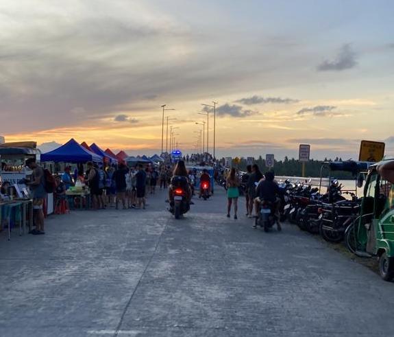 A road, with food stalls on the side, leading to a bridge on a sunset.