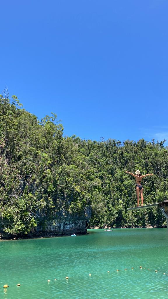 the sky and a greeny rocky hill and a blue -green lagoon with a woman on a wooden jump board at the right.